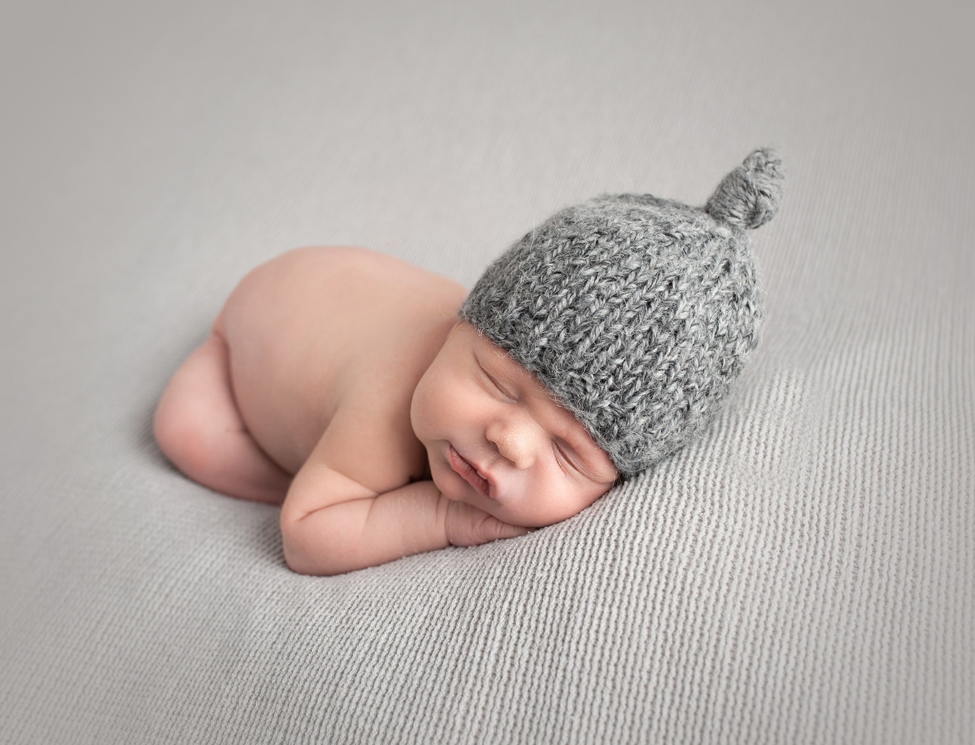 newborn baby wearing a hat posed asleep on grey blanket